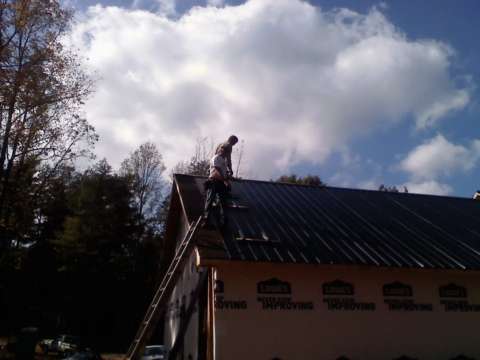 Roofer fastening dark metal panels high against a cloudy sky, from a Roof Inspection job in Moultonborough